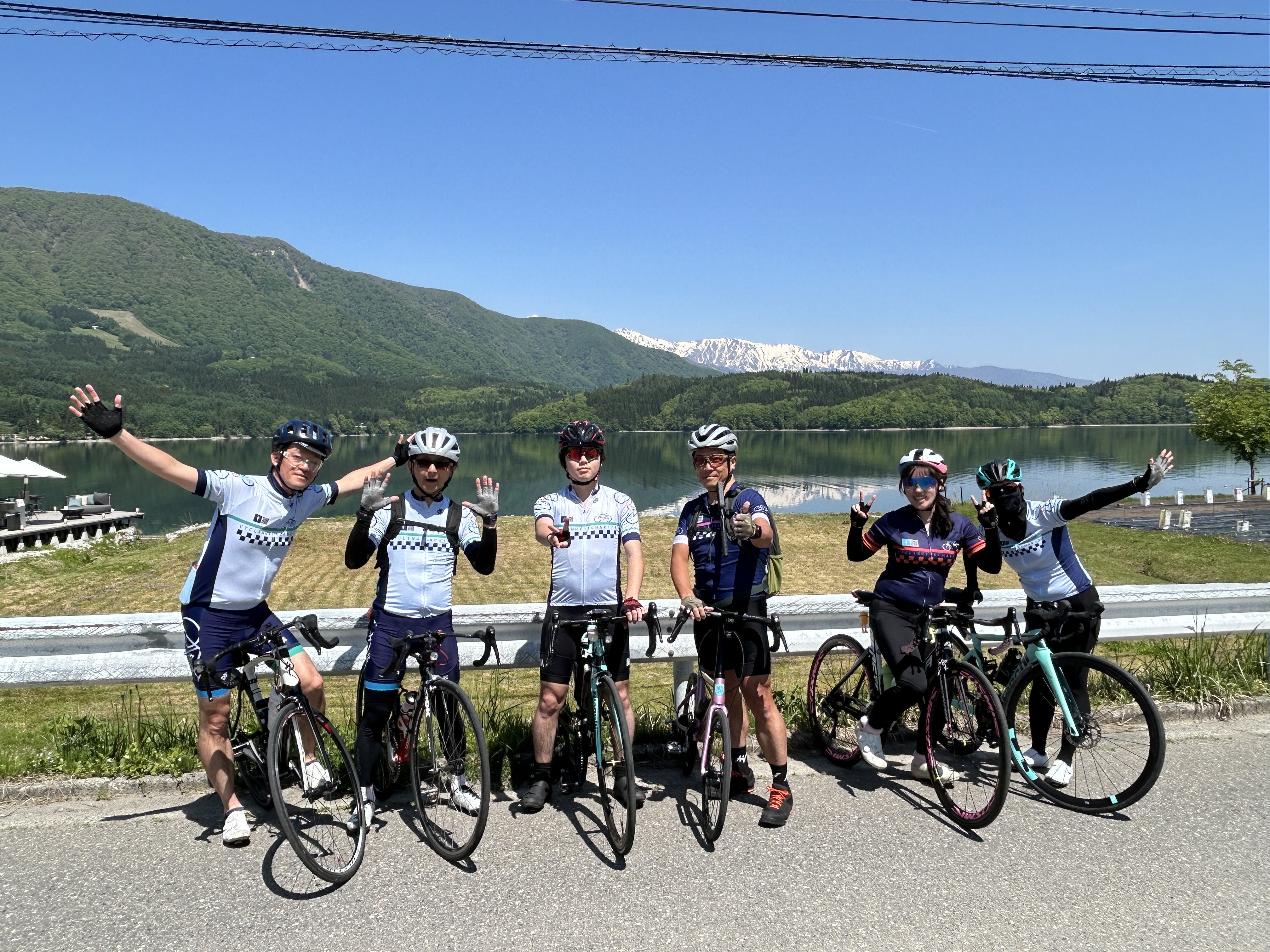 2026 North Alps — cyclists at lake with snow-capped Alps